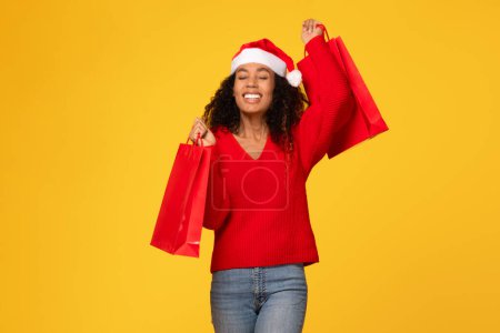 Photo for Joyful black woman donned in Santa hat, clutching red shopper bags, suggesting successful Christmas sales, standing against yellow background - Royalty Free Image