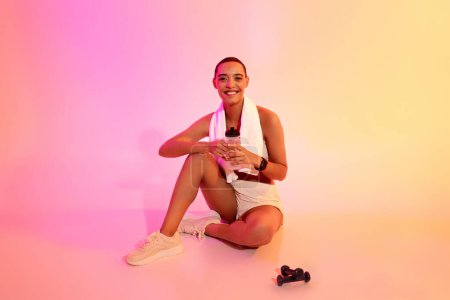 Téléchargez les photos : Joyeux jeune femme latine en tenue de sport assis avec serviette, bouteille d'eau, reposant de l'entraînement sur le sol sur fond de studio rose fluo. Pause de remise en forme, sport, soins de santé, perte de poids - en image libre de droit