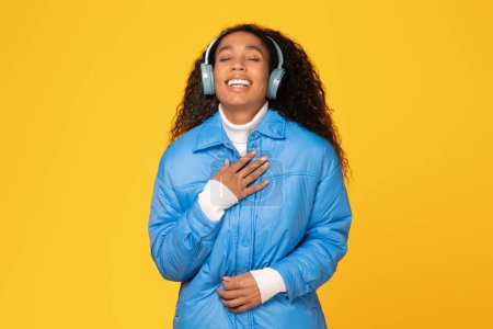 Téléchargez les photos : Jeune femme noire vêtue d'un manteau d'hiver se délecte de musique, chantant les yeux fermés, sur fond jaune ensoleillé, sentant la joie des fêtes - en image libre de droit