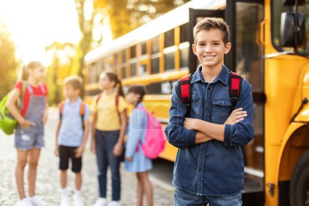 Foto de Sonriente niño preadolescente de pie con los brazos cruzados cerca del autobús escolar amarillo y mirando a la cámara, Grupo de sus compañeros de clase charlando y riendo en segundo plano, los niños listos para el nuevo día de aprendizaje - Imagen libre de derechos
