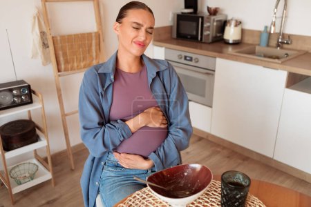 Foto de Mujer embarazada sostiene su vientre con expresión ligeramente incómoda, lo que ilustra la ocurrencia común de comer en exceso durante el embarazo - Imagen libre de derechos
