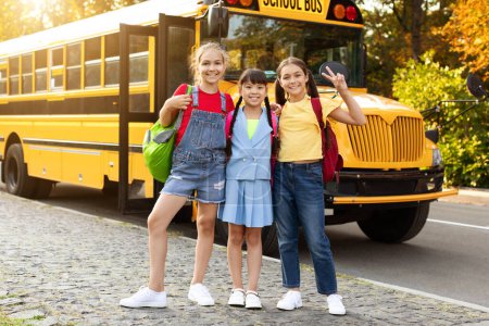 Foto de Retrato de tres niñas preadolescentes multiétnicas de pie afuera cerca del autobús escolar amarillo, lindos niños femeninos con mochilas abrazando y sonriendo a la cámara, listos para un nuevo día en la escuela, espacio para copiar - Imagen libre de derechos