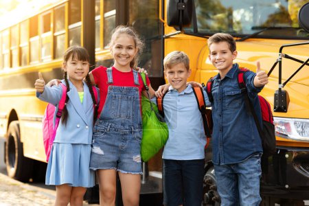 Foto de Alumnos multiétnicos felices de pie junto al autobús escolar amarillo y mostrando los pulgares hacia arriba en la cámara, grupo de niños alegres con mochilas abrazando y sonriendo, recomendando programas educativos - Imagen libre de derechos