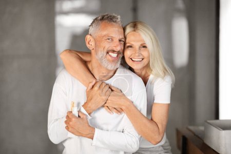 Téléchargez les photos : Portrait de heureux conjoints aînés embrassant l'intérieur de la salle de bains, aimer couple mature étreignant et souriant à la caméra, profiter de faire de la beauté du matin routine ensemble à la maison, Copier l'espace - en image libre de droit