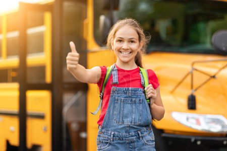 Foto de Alegre niño preadolescente de pie cerca del autobús escolar amarillo y mostrando el pulgar hacia arriba en la cámara, colegiala linda feliz usando mochila recomendando programas educativos, disfrutando de estudio, espacio de copia - Imagen libre de derechos