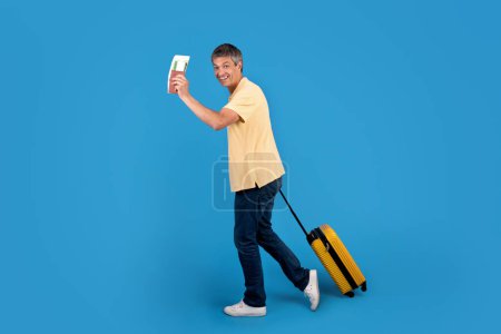Téléchargez les photos : Homme voyageur mature détient passeport et carte d'embarquement posant avec valise sur fond de studio bleu, l'excitation du voyage à venir et les promotions touristiques. Longueur totale - en image libre de droit