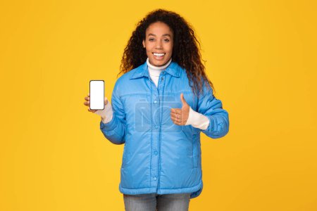 Foto de Sonriente dama negra vestida con acogedora chaqueta de invierno, mostrando con confianza la pantalla del teléfono inteligente en blanco con el gesto de pulgar hacia arriba, contra el fondo amarillo, maqueta - Imagen libre de derechos