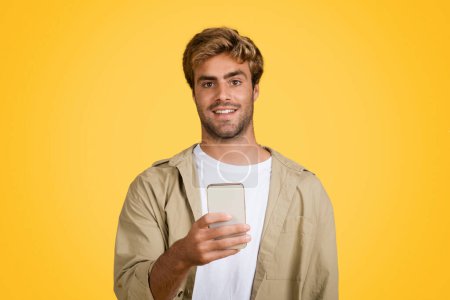 Téléchargez les photos : Jeune et élégant homme européen se tient avec confiance, téléphone portable à la main, gars sourit chaudement à la caméra sur fond jaune distinct - en image libre de droit