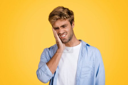 Téléchargez les photos : Jeune homme européen met en valeur l'inconfort évident, toucher sa joue en raison de maux de dents, représentation claire de la douleur dentaire sur fond de studio jaune vif - en image libre de droit