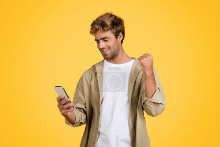 Foto de Joven hombre europeo con estilo irradia alegría mientras sostiene el teléfono celular, apretando los puños en la emoción, posicionado contra el fondo del estudio de color amarillo brillante - Imagen libre de derechos