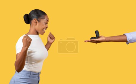 Foto de Mujer negra emocionada con sonrisa brillante mirando la caja del anillo abierto presentada por la mano masculina contra el fondo amarillo vibrante, expresando felicidad y sorpresa - Imagen libre de derechos