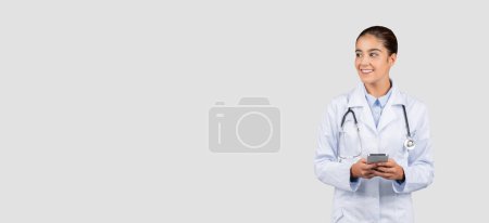 Foto de Sonriente doctora milenaria europea en bata blanca, con estetoscopio, charlando por teléfono, mira el espacio de copia, aislado en el fondo gris del estudio. Medicina, atención médica aplicación en línea - Imagen libre de derechos