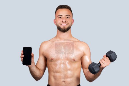 Photo for Glad sweat strong muscular millennial european man athlete with beard, naked torso make exercises with dumbbell, strength workout, show phone with empty screen, isolated on gray studio background - Royalty Free Image