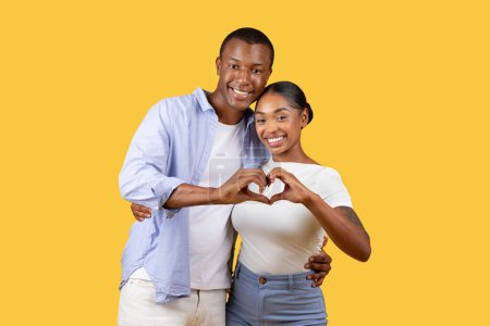 Foto de Cariñosa joven pareja negra sonriendo y creando forma de corazón junto con sus manos sobre alegre fondo amarillo, simbolizando el amor y la conexión - Imagen libre de derechos