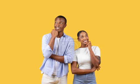 Téléchargez les photos : Jeune homme noir réfléchi et femme avec les mains sur le menton regardant vers le haut, exprimant curiosité et contemplation sur fond jaune solide - en image libre de droit