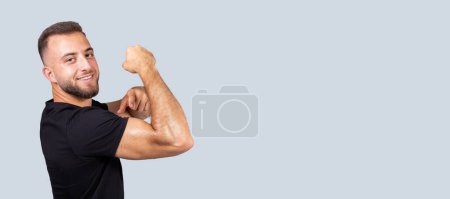 Téléchargez les photos : Joyeux confiant fort millénariste européen homme avec barbe en vêtements de sport doigt pointeur aux biceps musculaires sur la main, isolé sur fond gris. Soins du corps, mode de vie sportif et recommandation d'entraînement - en image libre de droit