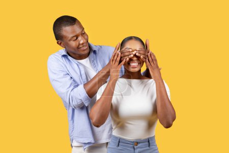 Photo for Happy black man surprises delighted woman by covering her eyes with his hands, both smiling in anticipation of surprise on yellow background - Royalty Free Image