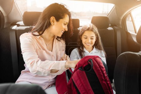 Foto de Dentro del coche, la madre sonriente interactúa amorosamente con su hija, ya que ambos se centran en la mochila de la escuela roja, creando una atmósfera cálida y conectada. - Imagen libre de derechos