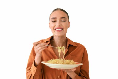 Photo for Delightful young lady in orange blouse holds plate of delicious spaghetti, smiling with eyes closed as shes about to take delicious bite on white backdrop - Royalty Free Image