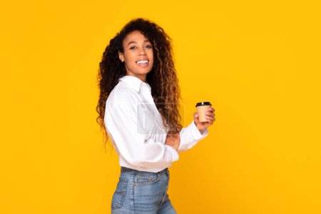 Téléchargez les photos : Jeune femme afro-américaine aux cheveux bouclés tenant une tasse de café en papier, souriant à la caméra debout sur fond de studio jaune, portant une chemise blanche et un jean. Profitez de votre concept de pause - en image libre de droit