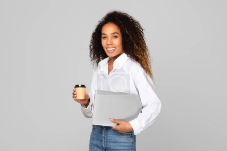 Foto de Mujer afroamericana sonriendo sosteniendo su computadora portátil, de pie con una taza de café de papel, en un estudio sobre fondo gris. Tecnologías digitales, concepto de carrera independiente y en línea - Imagen libre de derechos