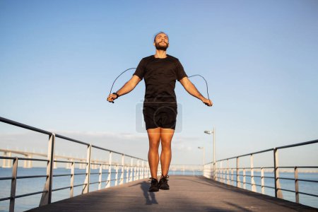 Photo for Full length of young sportsman with jumping rope exercising at sea pier outdoors, copy space. Millennial european man in fitwear doing cardio workout or aerobic exercises outside - Royalty Free Image