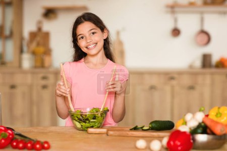 Photo for Happy european young girl in stirs bowl of fresh salad in a well-lit, spacious kitchen with vegetables on the counter, smiling at camera, free space - Royalty Free Image