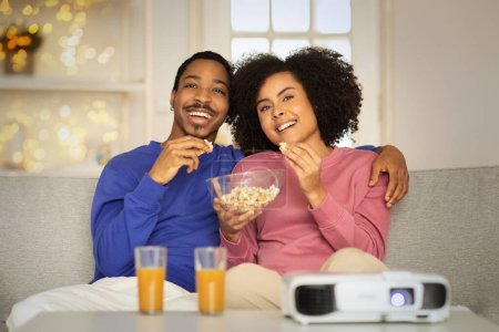 Photo for Cheerful black young couple cuddling on the sofa, enjoying a movie via projector in modern living room at home, snacking on popcorn. Perfect weekend relaxation together - Royalty Free Image
