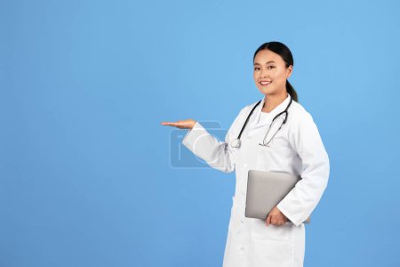 Sonriente asiático médico femenino en uniforme celebración de la computadora portátil y señalando a un lado con la palma, terapeuta amigable señora publicidad producto o clínica, doc mujer posando aislado en azul estudio fondo