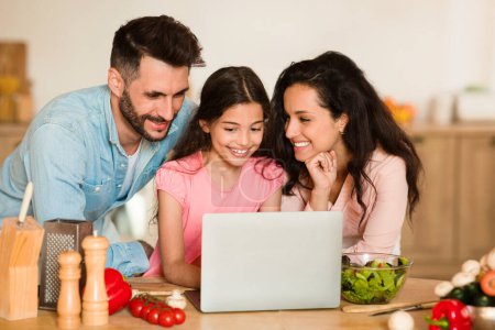 Téléchargez les photos : Famille européenne heureuse avec jeune fille sourit tout en regardant ordinateur portable dans la cuisine remplie d'ingrédients alimentaires sains, cuisiner ensemble et partager le moment - en image libre de droit