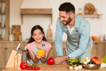 Téléchargez les photos : Papa souriant et sa jeune fille profiter du temps ensemble dans la cuisine, avec la fille tenant poivron jaune, prêt pour une leçon de cuisine saine - en image libre de droit