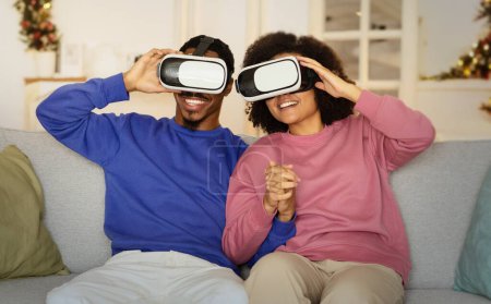Photo for Virtual Reality Fun. Young black husband and wife engrossed in online simulation game via VR headsets, sitting on sofa at home interior. Modern tech weekend leisure concept - Royalty Free Image