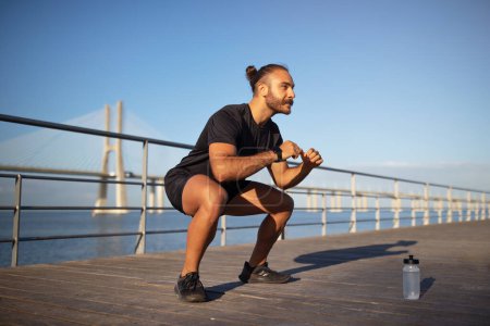 Photo for Positive young caucasian man enjoys outdoor fitness workout while squats at sea pier. Athletic guy having sports on weekend morning. Active lifestyle and body care, weight loss - Royalty Free Image
