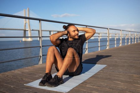 Photo for Cheerful sporty guy exercising on pier outside next to the sea, lying on yoga mat and doing abs crunches, working on abdominal muscles and smiling looking aside. Outdoor fitness concept - Royalty Free Image
