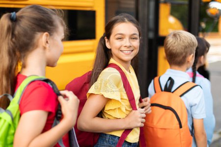 Foto de Retrato de niña sonriente con mochila abordaje autobús escolar amarillo con compañeros de clase y mirando a la cámara, Grupo de niños que entran en el vehículo después de lecciones, listos para el viaje juntos, Primer plano - Imagen libre de derechos