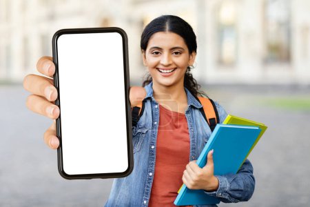 Photo for Cheerful indian woman student showing smartphone with white blank screen, posing at campus, copy space, mockup. Positive lady with backpack and wireless headset recommending educational app - Royalty Free Image