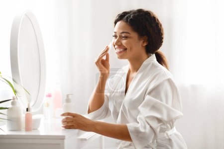 Téléchargez les photos : Soin quotidien. Jeune femme noire nettoyant la peau avec coton tampon et le lait de démaquillage tout en étant assis près du miroir à la maison, souriant afro-américaine faisant la routine de beauté domestique, espace libre - en image libre de droit