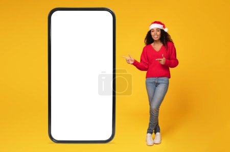 Téléchargez les photos : Joyeuse dame afro-américaine au chapeau de Père Noël pointant vers un grand téléphone avec écran blanc blanc sur fond jaune, maquette, endroit pour l'offre de Noël - en image libre de droit