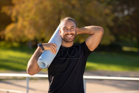 Téléchargez les photos : Jeune homme sportif souriant avec confiance à la caméra tout en tenant tapis de fitness dans un parc serein. Le gars est prêt pour l'entraînement de yoga. Fitness extérieur et rituels matinaux sains - en image libre de droit