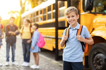 Foto de Sonriente niño preadolescente con mochila posando junto al autobús escolar amarillo mientras sus compañeros de clase charlan sobre el fondo, lindo niño masculino listo para el estudio, disfrutando de la educación y la vida escolar, enfoque selectivo - Imagen libre de derechos