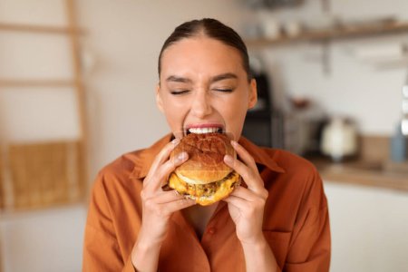 Téléchargez les photos : Femme déterminée en chemise prend une morsure enthousiaste de hamburger appétissant, tandis que dans la cuisine de la maison chaleureusement éclairée avec des appareils modernes visibles derrière - en image libre de droit