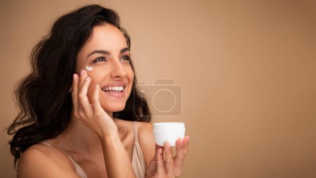 Photo for Cheerful long-haired brunette millennial woman with smile on her face using facial eye cream for young smooth glowing skin, isolated on beige studio background, closeup shot, copy space - Royalty Free Image