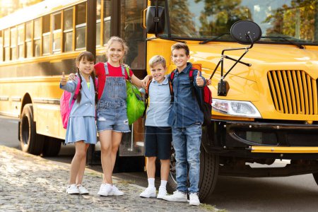 Téléchargez les photos : Groupe d'enfants heureux montrant pouce vers le haut tout en se tenant près du bus scolaire jaune à l'extérieur, souriants enfants avec des sacs à dos prêts pour l'étude, posant à l'extérieur avant d'aller aux leçons, bénéficiant de l'éducation - en image libre de droit