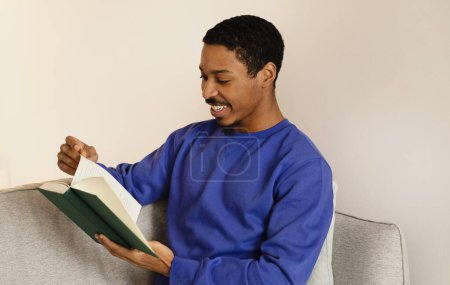Foto de Joven afroamericano absorto en la literatura, leyendo el libro atentamente mientras está sentado en un sofá acogedor en la sala de estar moderna, personificando la relajación y el intelecto en el fin de semana - Imagen libre de derechos