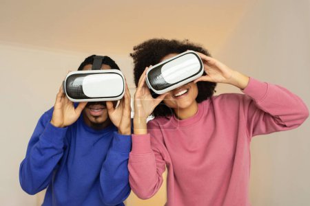 Téléchargez les photos : Couple afro-américain posant avec des lunettes VR, profitant d'une séance de jeu futuriste, posant ludique contre le mur du studio, regardant la caméra à travers des lunettes. Plaisir numérique augmenté - en image libre de droit