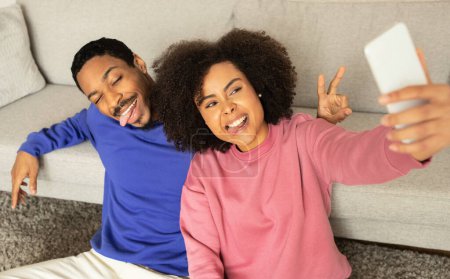 Téléchargez les photos : Jeune couple noir capturant moment avec selfie sur leur smartphone, se détendre dans l'intérieur du salon domestique, posant ensemble et s'amusant. Romance moderne et gadgets - en image libre de droit