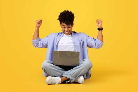 Téléchargez les photos : Jeune homme noir exubérant assis jambes croisées, les bras levés dans la victoire, tout en regardant ordinateur portable sur fond jaune vif - en image libre de droit