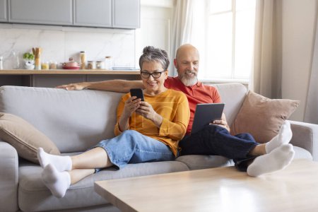 Téléchargez les photos : Conjoints aînés heureux en utilisant des gadgets modernes tout en se relaxant sur le canapé à la maison, sourire homme et femme âgés naviguant sur Internet ou faire du shopping en ligne tout en se reposant dans le salon intérieur, Copier l'espace - en image libre de droit