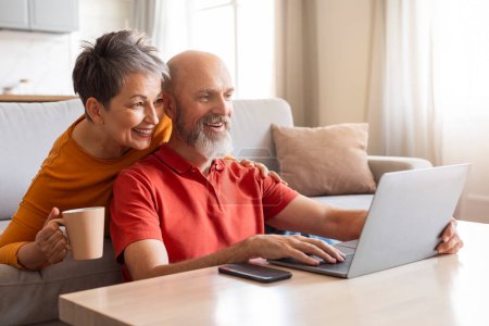 Photo for Weekend Pastime. Happy Senior Couple Resting With Laptop And Coffee In Living Room Interior, Cheerful Mature Spouses Using Computer Together At Home And Enjoying Hot Drinks, Free Space - Royalty Free Image