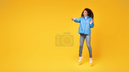 Téléchargez les photos : Jeune femme noire veste d'hiver saute haut, pointant vers un espace vide sur fond jaune, plein de joie de vacances et d'humeur vibrante, pleine longueur, panorama - en image libre de droit